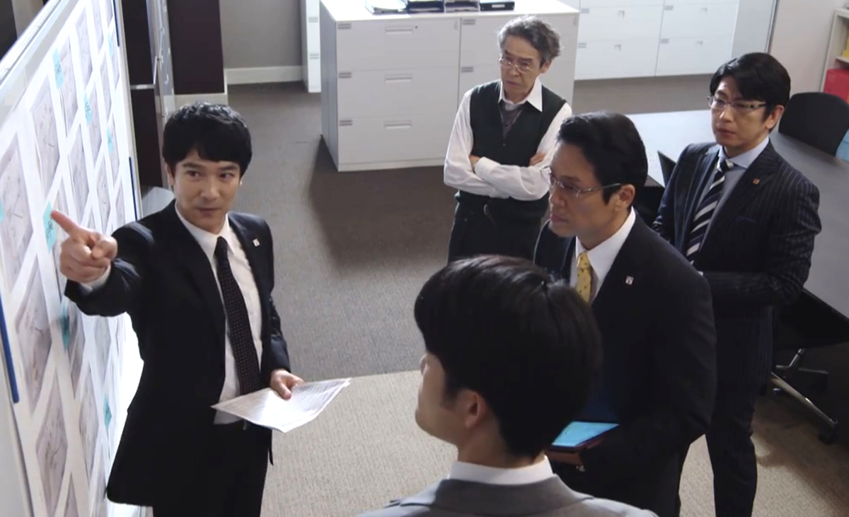 Screencap from Hanzawa Naoki, showing Naoki Hanzawa pointing out something on a board to his colleagues